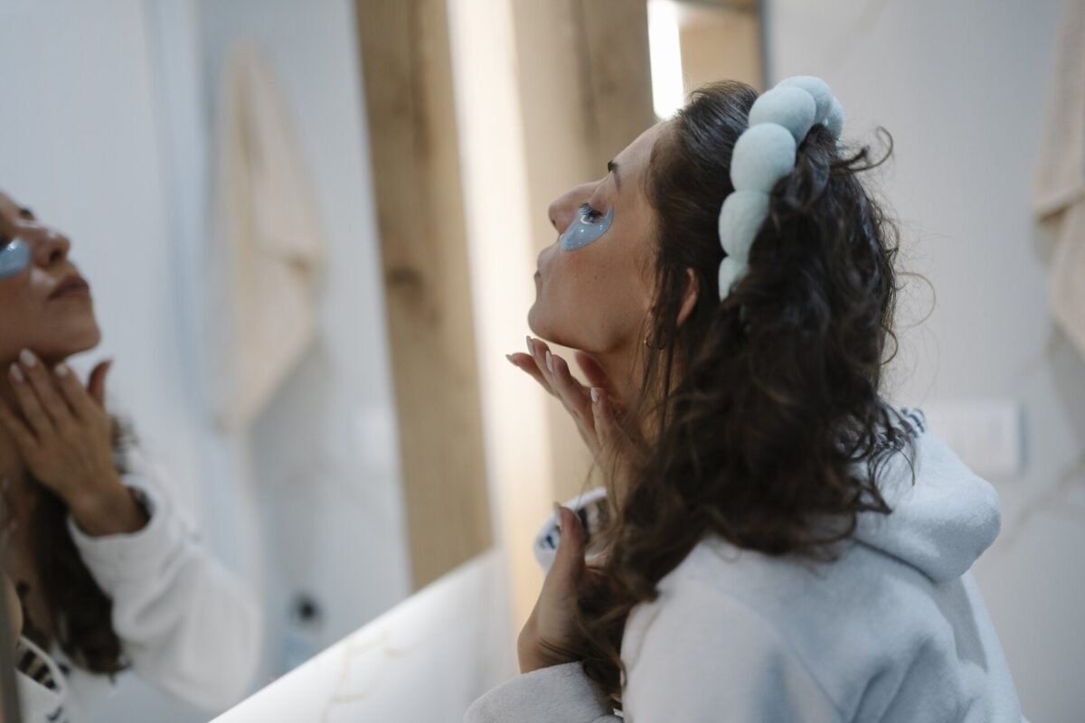 A woman is standing in front of a mirror in a bathroom. She is using a skincare product on her face while dressed in comfortable clothes. The setting
