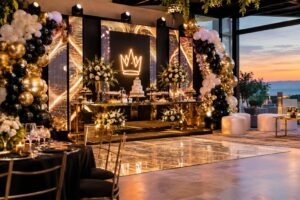 Ambiente decorado para festa de 15 anos com mesa espelhada, bolo, arranjos florais brancos, painéis de LED em tons dourados e uma iluminação quente. Na decoração há arcos de balões em cores dourado, preto e branco, cadeiras pretas com detalhes dourados e um pôr do sol ao fundo.