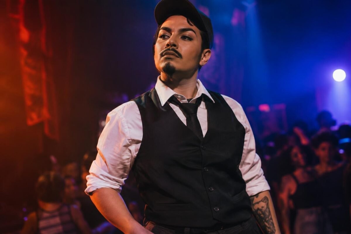 Artista drag king em palco com figurino masculino composto por colete preto, camisa branca com mangas dobradas, gravata e boné, olhando para o lado com expressão confiante, em ambiente iluminado por luzes coloridas e público ao fundo.